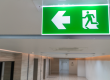 Illuminated green fire exit sign with a left-pointing arrow above a corridor in a building