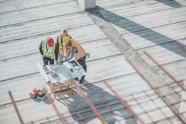 Construction team reviewing plans on site Three construction professionals in hi-vis jackets reviewing building plans on an open construction site, viewed from above.