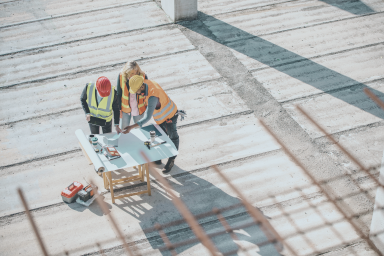 Construction team reviewing plans on site Three construction professionals in hi-vis jackets reviewing building plans on an open construction site, viewed from above.